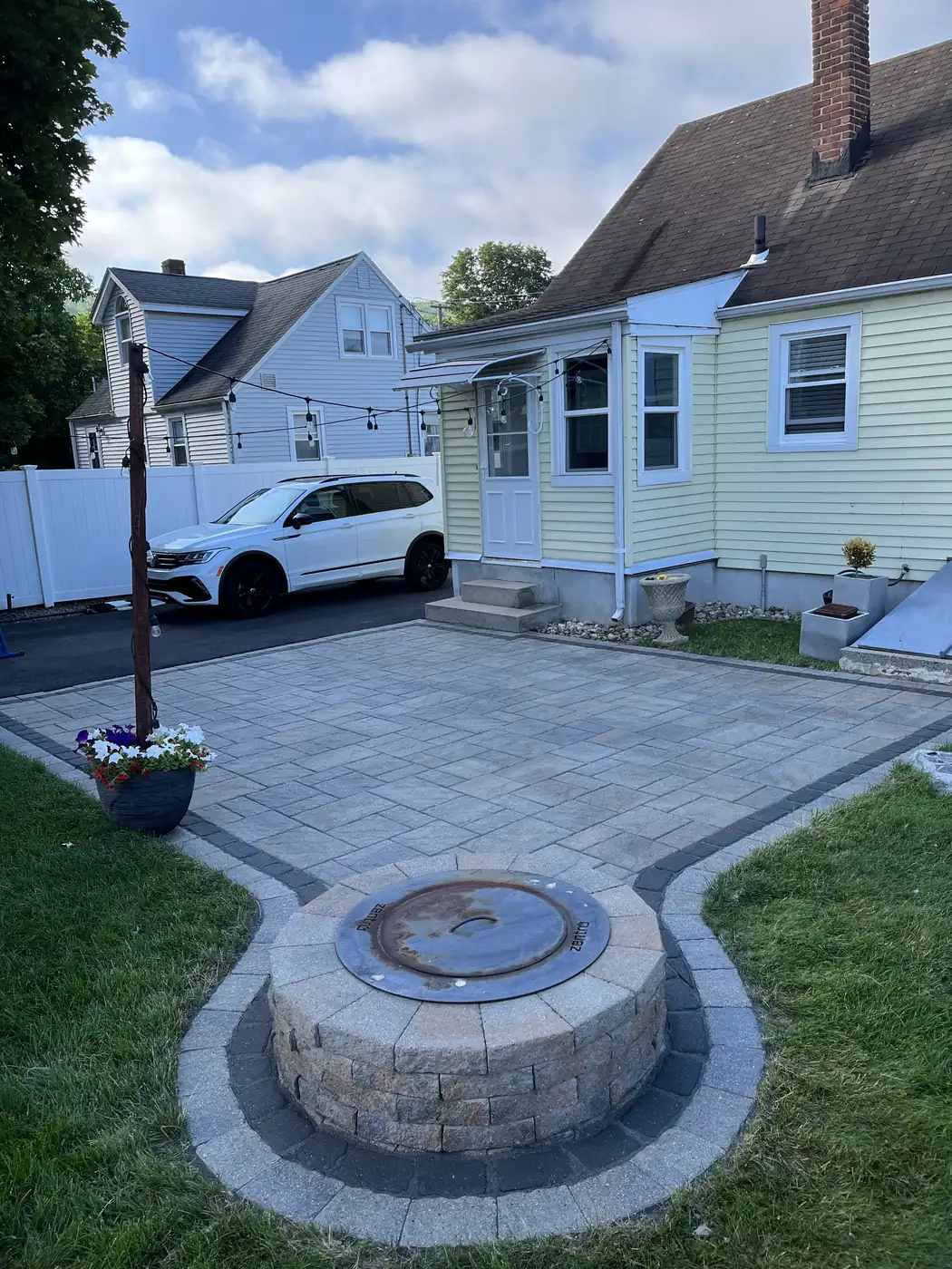 Keyhole paver patio with a round fire pit and charcoal border behind a cape home in Plainville, CT